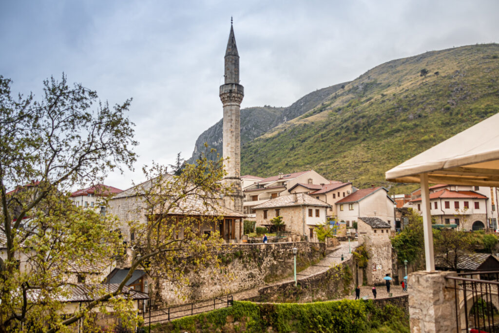 A tall minaret rises over the rooftops of Mostar, green hills in the background