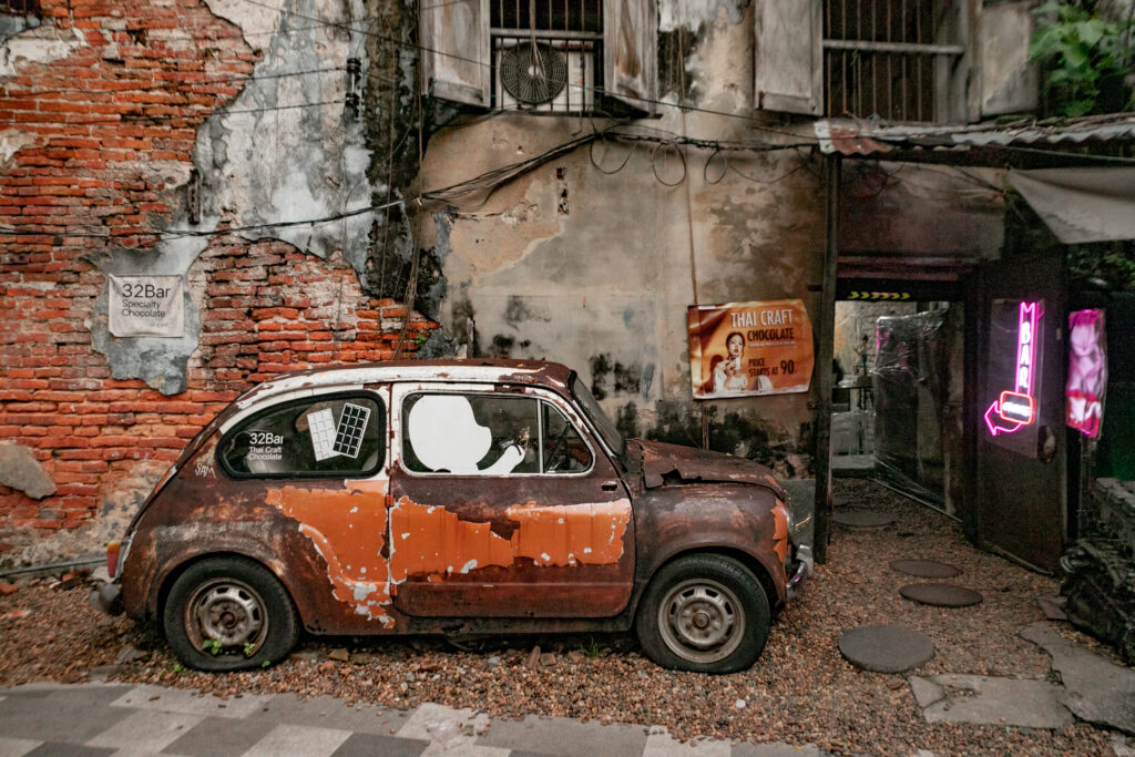 A rusty old car placed outside a cafe with a neon pink sign