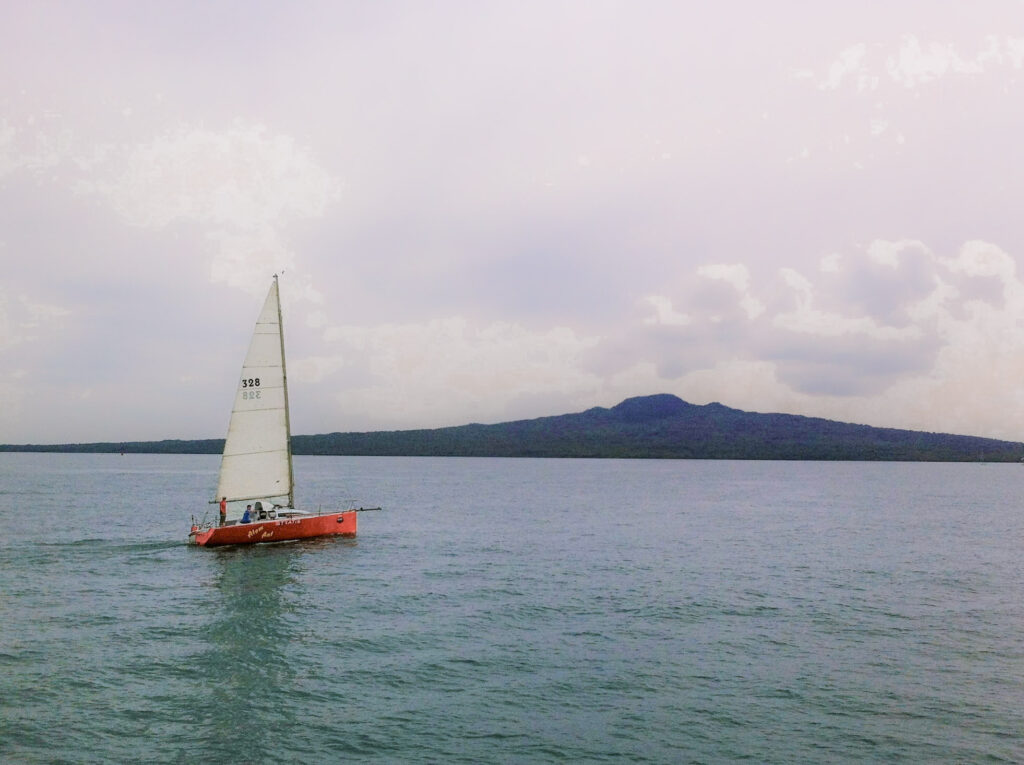 A red yacht with white sails on the water in front of Rangitoto Island on the horizon