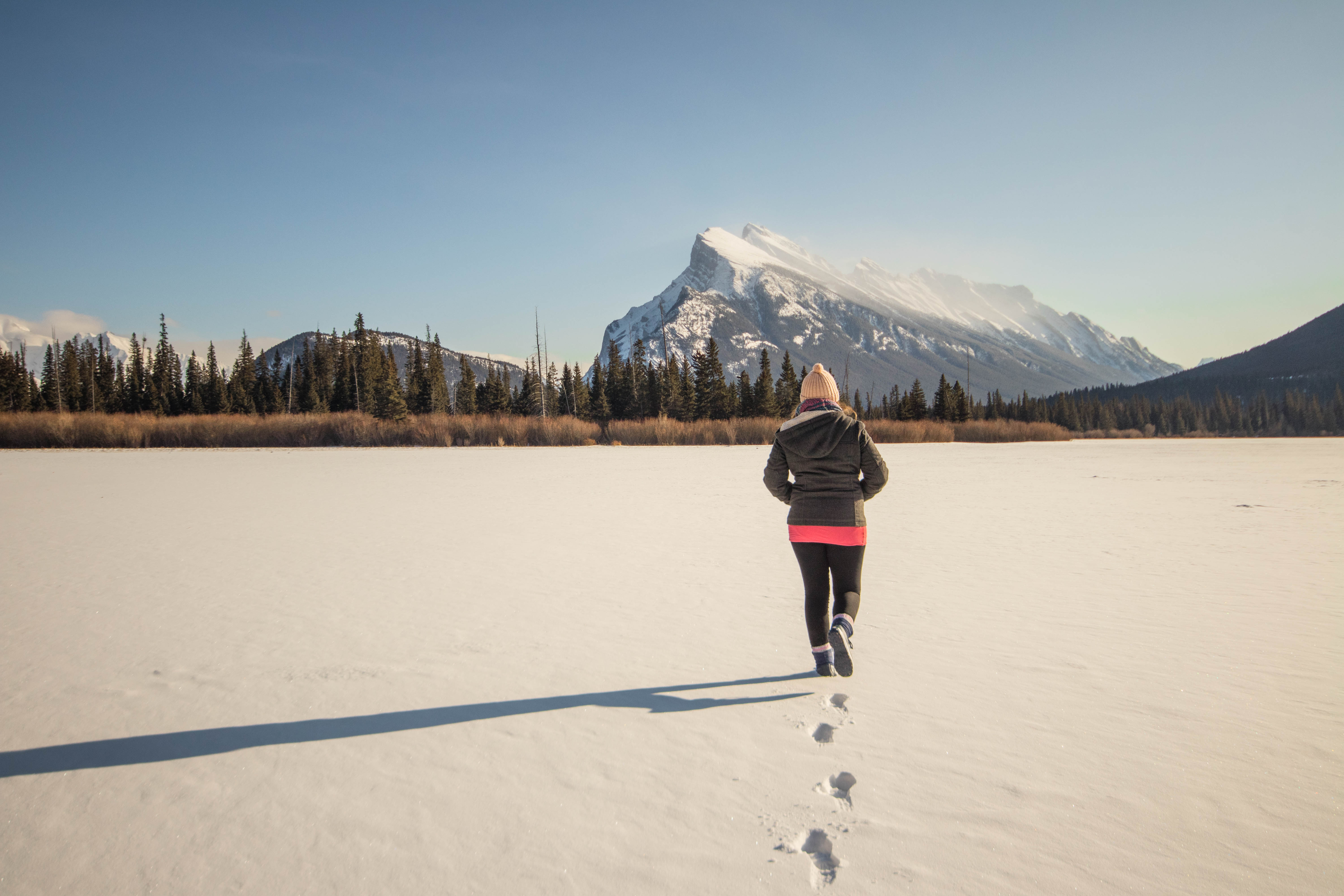 A girl walks away from the camera over a snow covered lake, a distinctive mountain in the bckground
