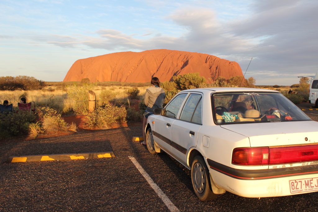 A white car parked in a car park with Uluru in the background