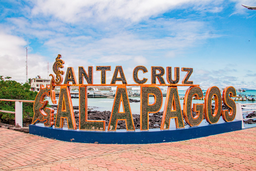 A large sign with the words Santa Cruz Galapagos in front of a rocky beach