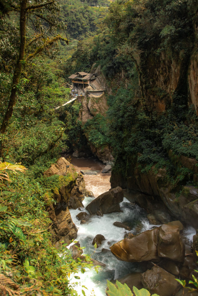 A river rushes over boulders in a narrow canyon, a building and swing bridge over the water