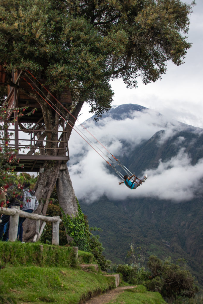 A person swings from a treehouse with a cloud-covered mountain in the background. The Swing at the End of the World is one of the most popular attractions in Banos and a must-do in any Ecuador itinerary