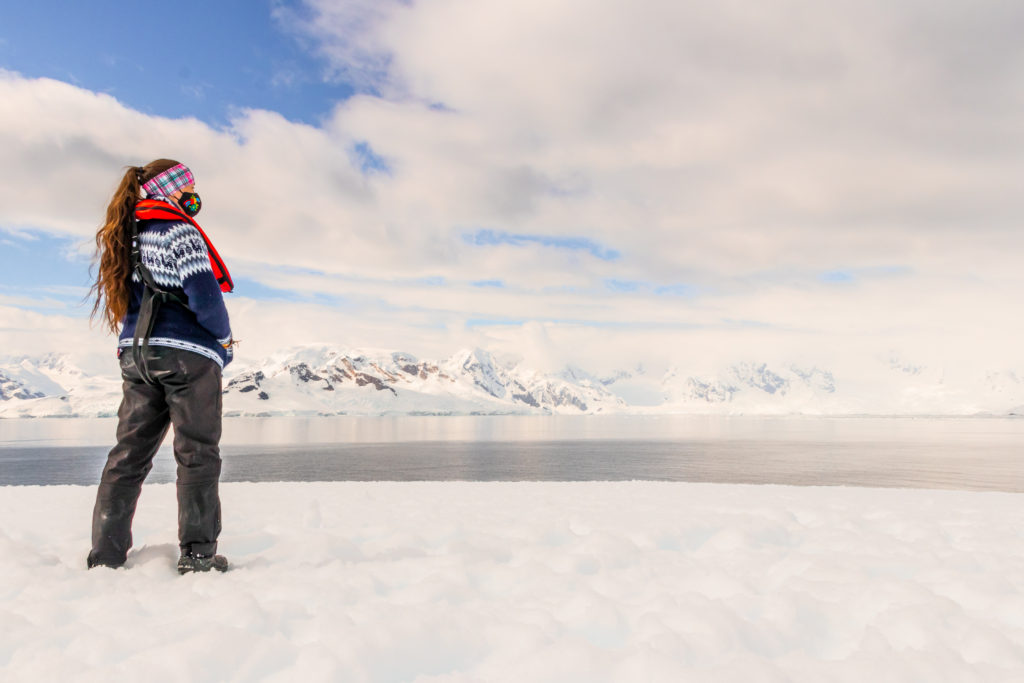 A figure stands on the right, looking out at a bay surrounded by snowy mountains - a backpacker in Antarctica, living the dream!