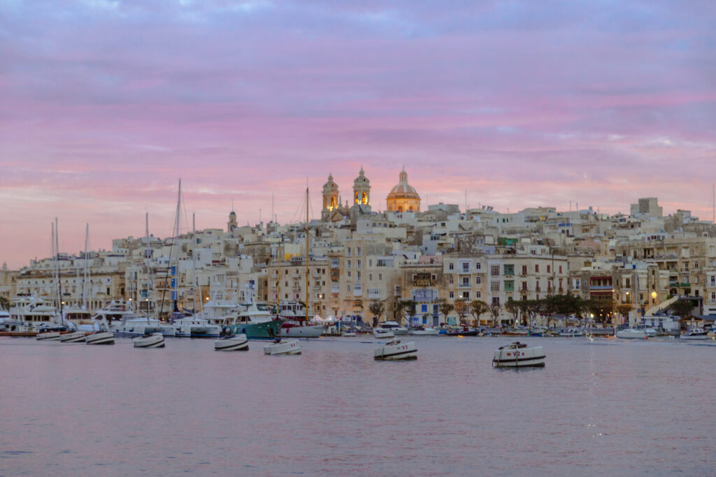 Purple skies over Senglea at sunset, with boats in the water and a church silhouetted on the top of the hill, Malta