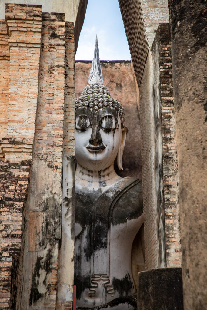 A large Buddha statue looks out through a narrow gap in a Sukhothai temple